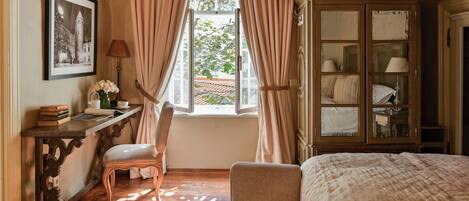 A bedroom with a bed, a desk with books and a lamp, a chair, and a window with curtains.