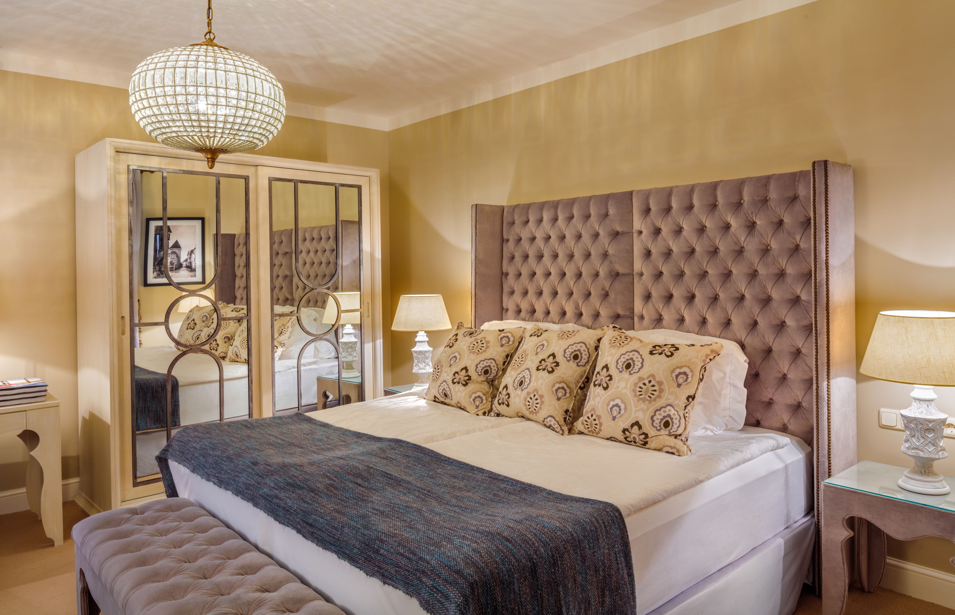 Une chambre à coucher avec un grand lit, une tête de lit capitonnée, un miroir, des tables de chevet et un lustre.