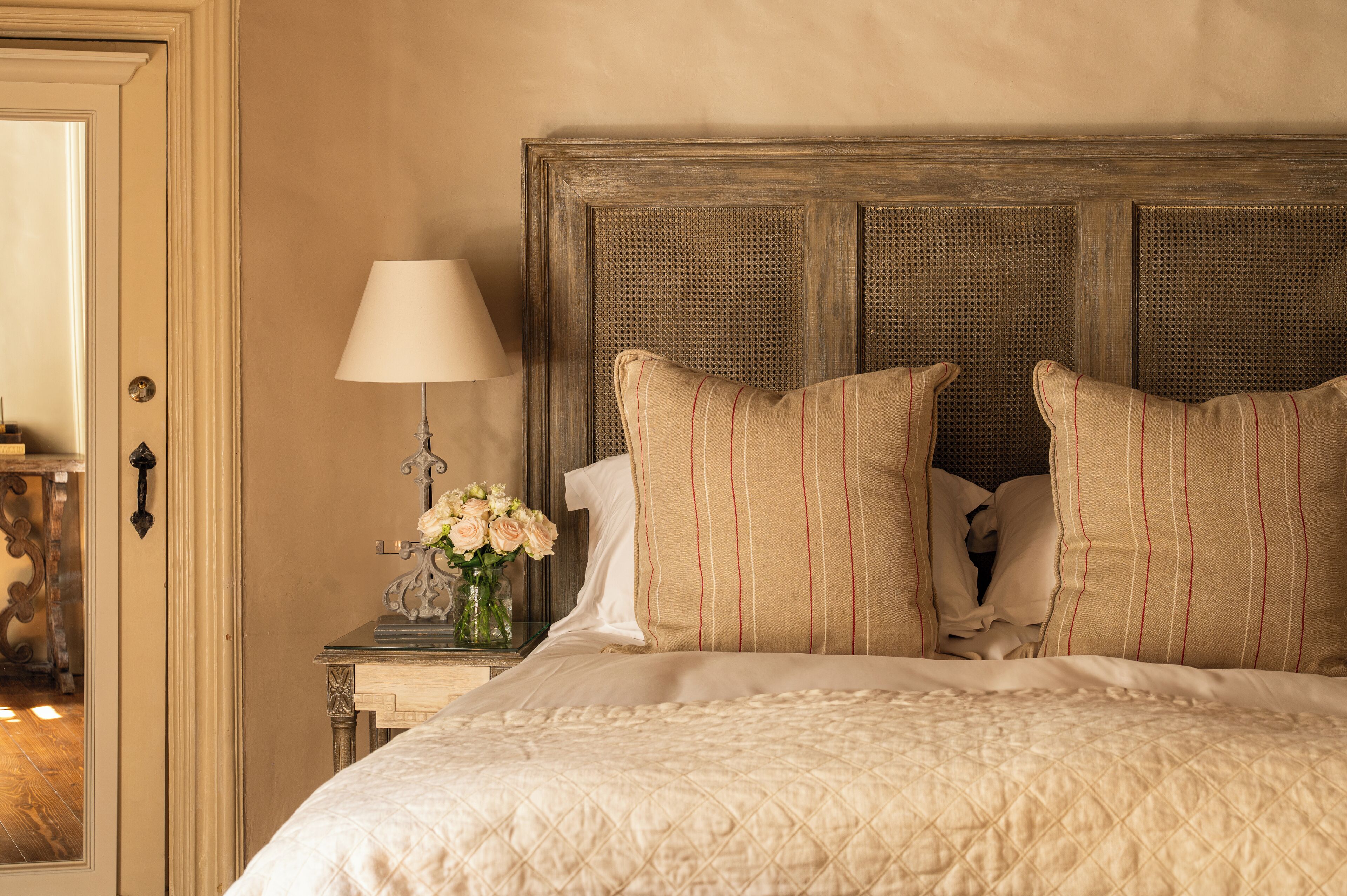 Una cama bien hecha con un cubrecolchón acolchado, una mesita de noche con una lámpara y un florero, y un cabecero de madera.