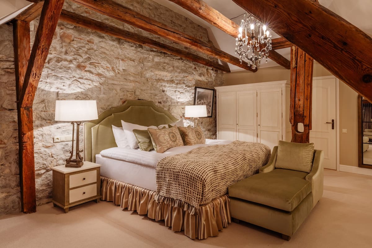 A bedroom with a stone wall, wooden beams, a bed with a green headboard, a green armchair, and a chandelier.