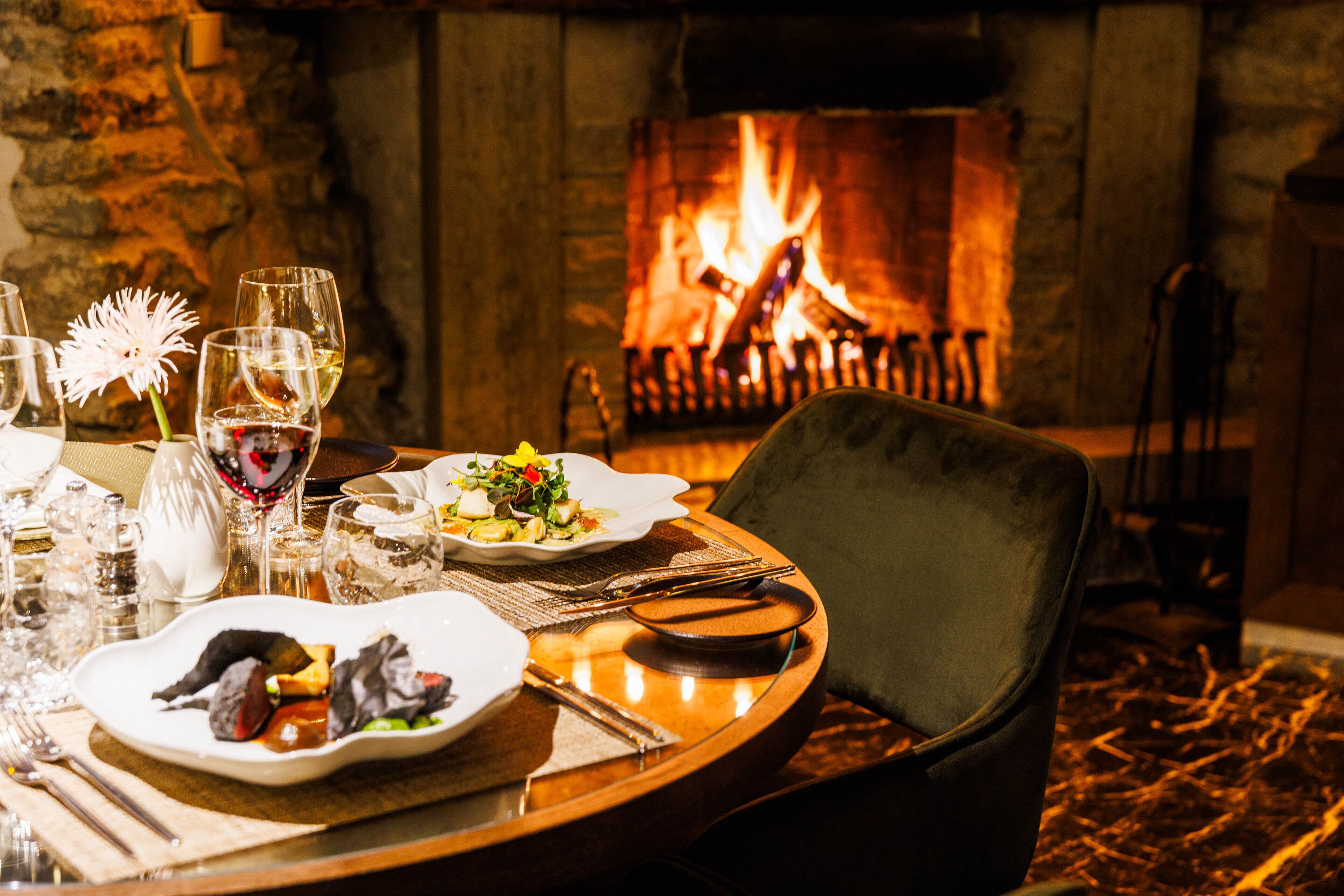 Una mesa de comedor dispuesta con platos, vasos y cubiertos, con una chimenea al fondo.