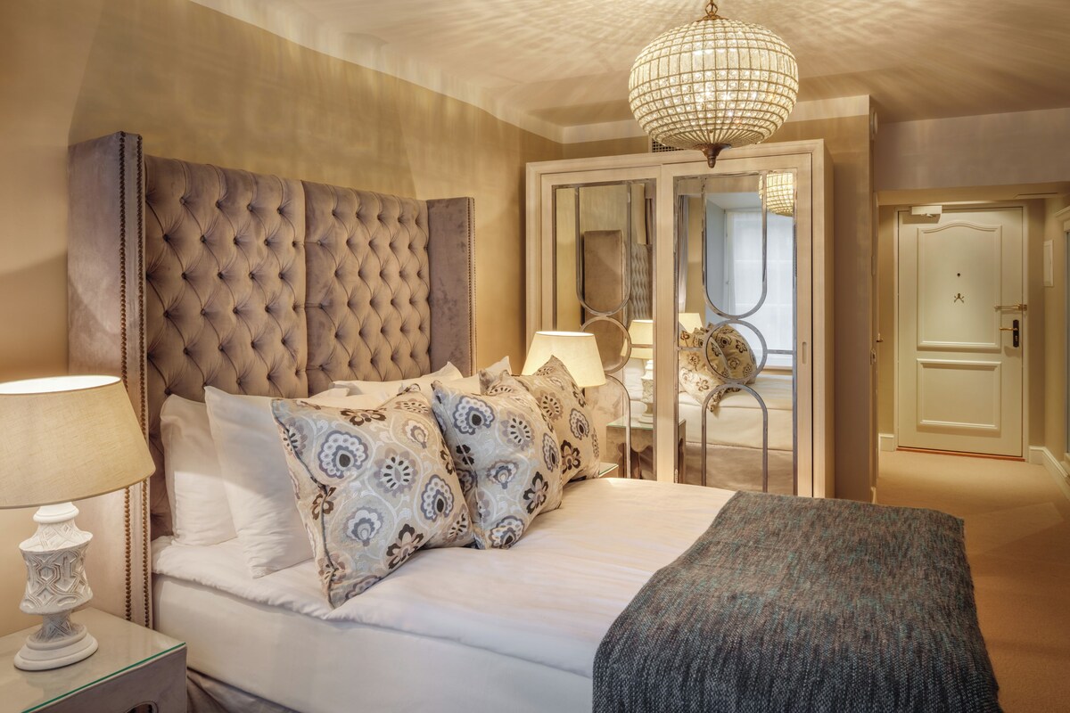 A bedroom with a tufted headboard, patterned pillows, a bedside table with a lamp, and a large mirror.