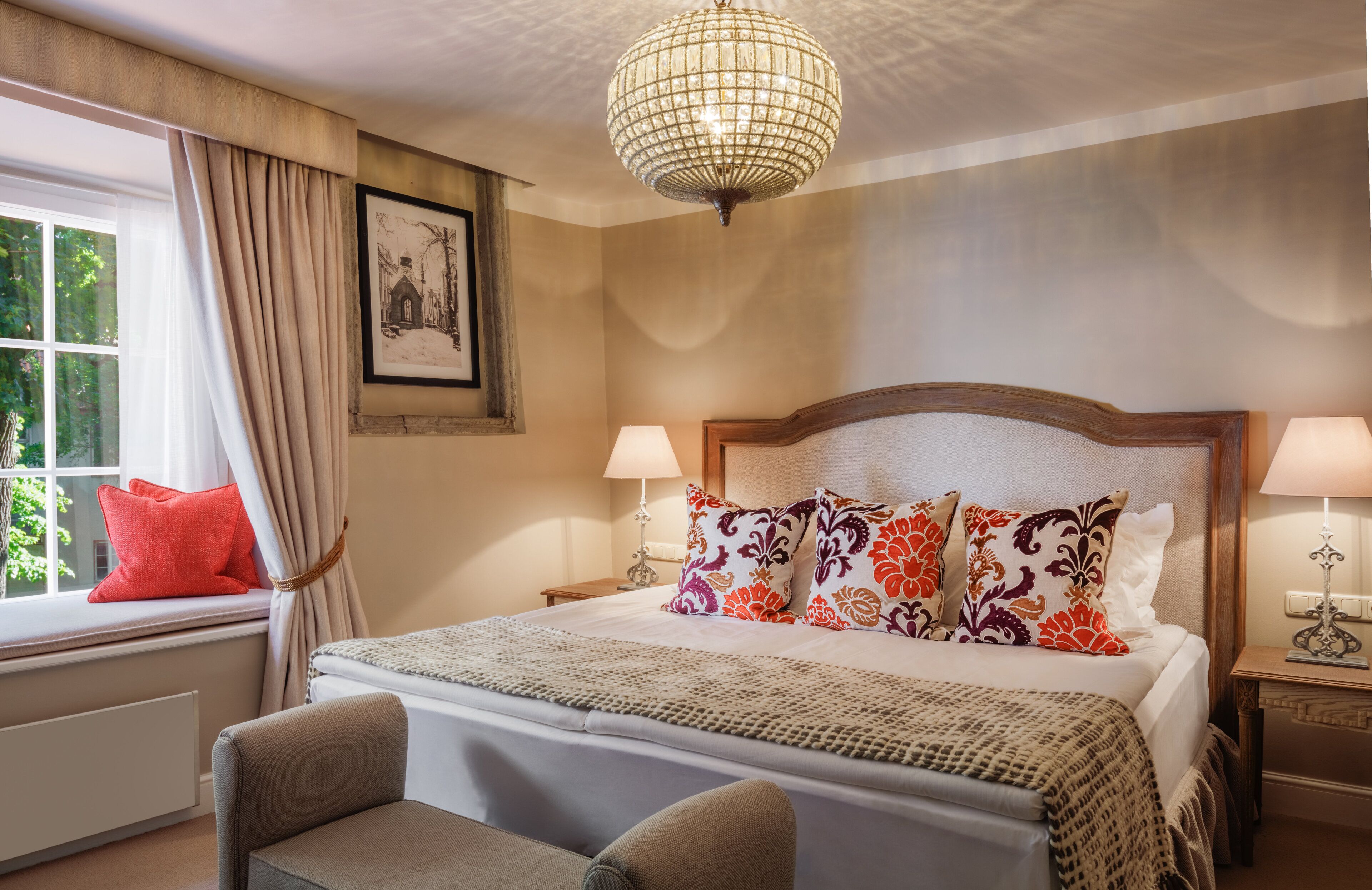 A neatly arranged hotel room with a large bed, two bedside tables, a window with curtains, a chandelier, and a framed picture on the wall.