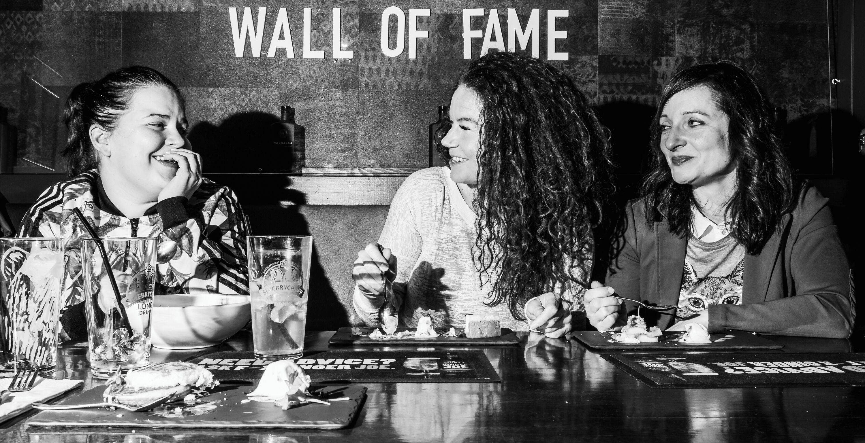 Tres personas sentadas en una mesa con vasos y un plato de comida, con un cartel que dice 
