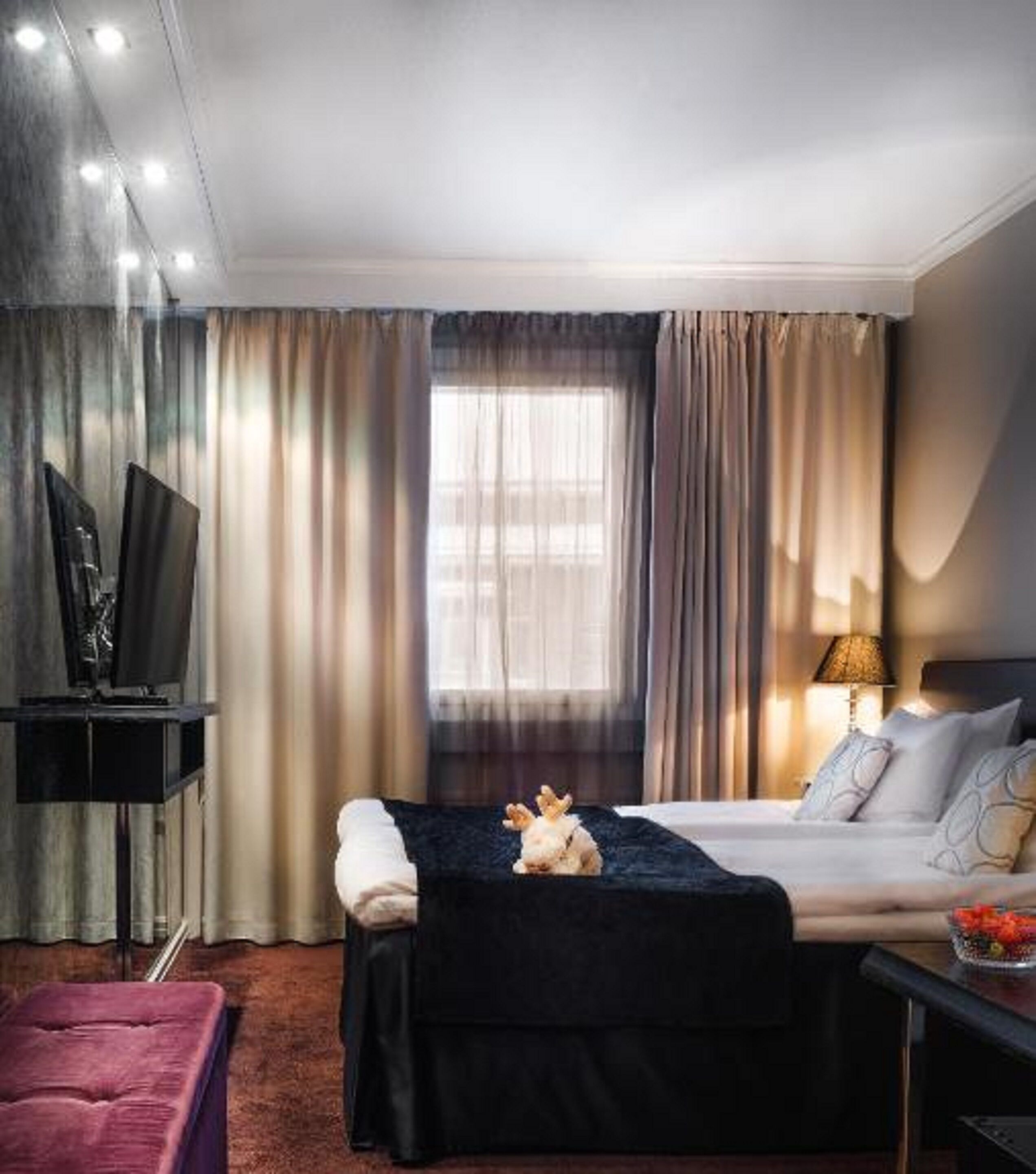 Quarto de hotel com uma cama, televisão, escrivaninha e janela com cortinas.