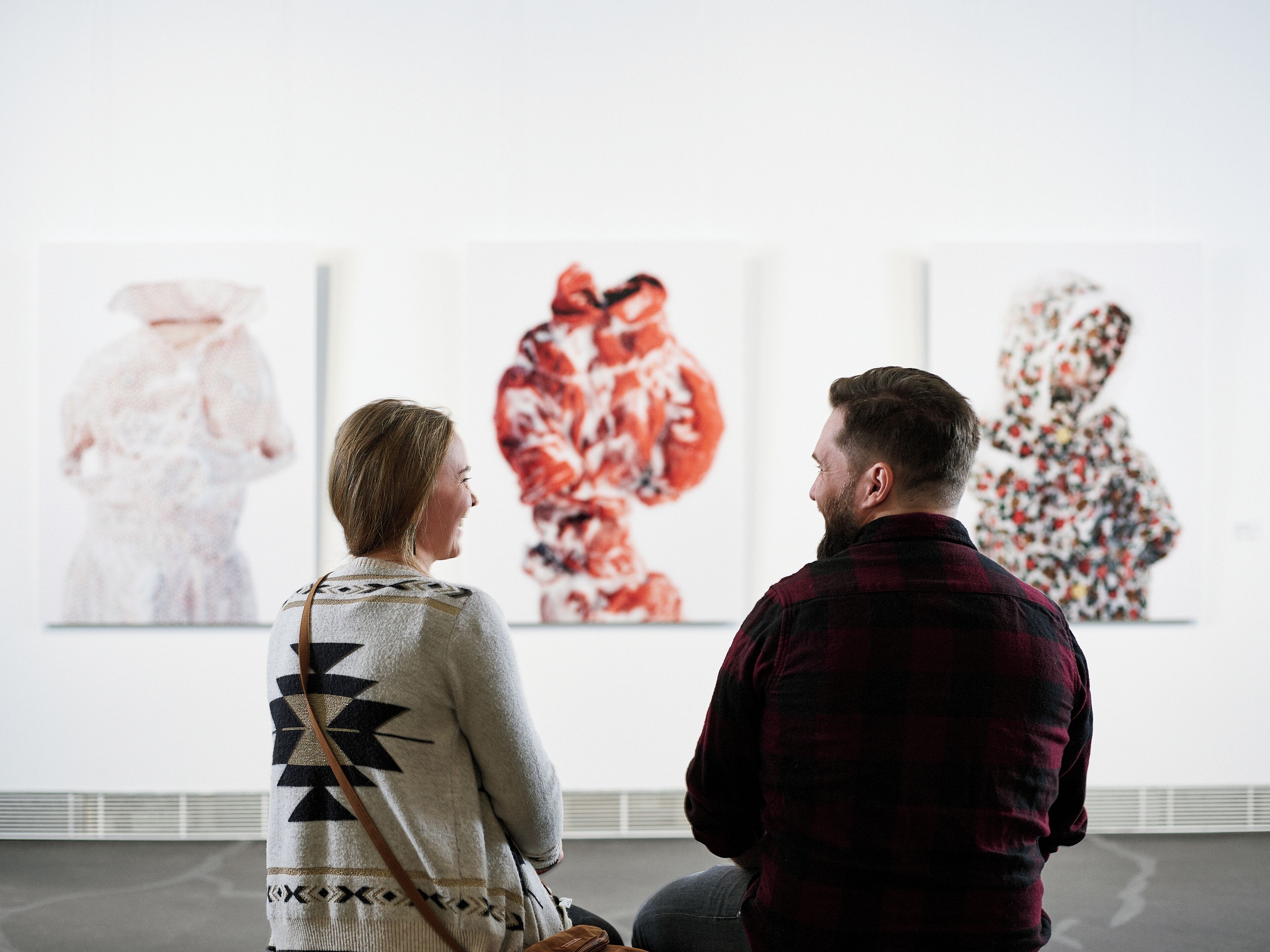 Two people observing artworks in a gallery with white walls and a simple floor layout.