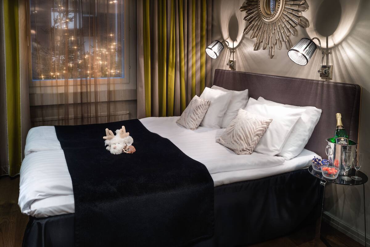 A hotel room with a neatly made bed, a nightstand with a bottle of champagne, and a view of the city through sheer curtains.