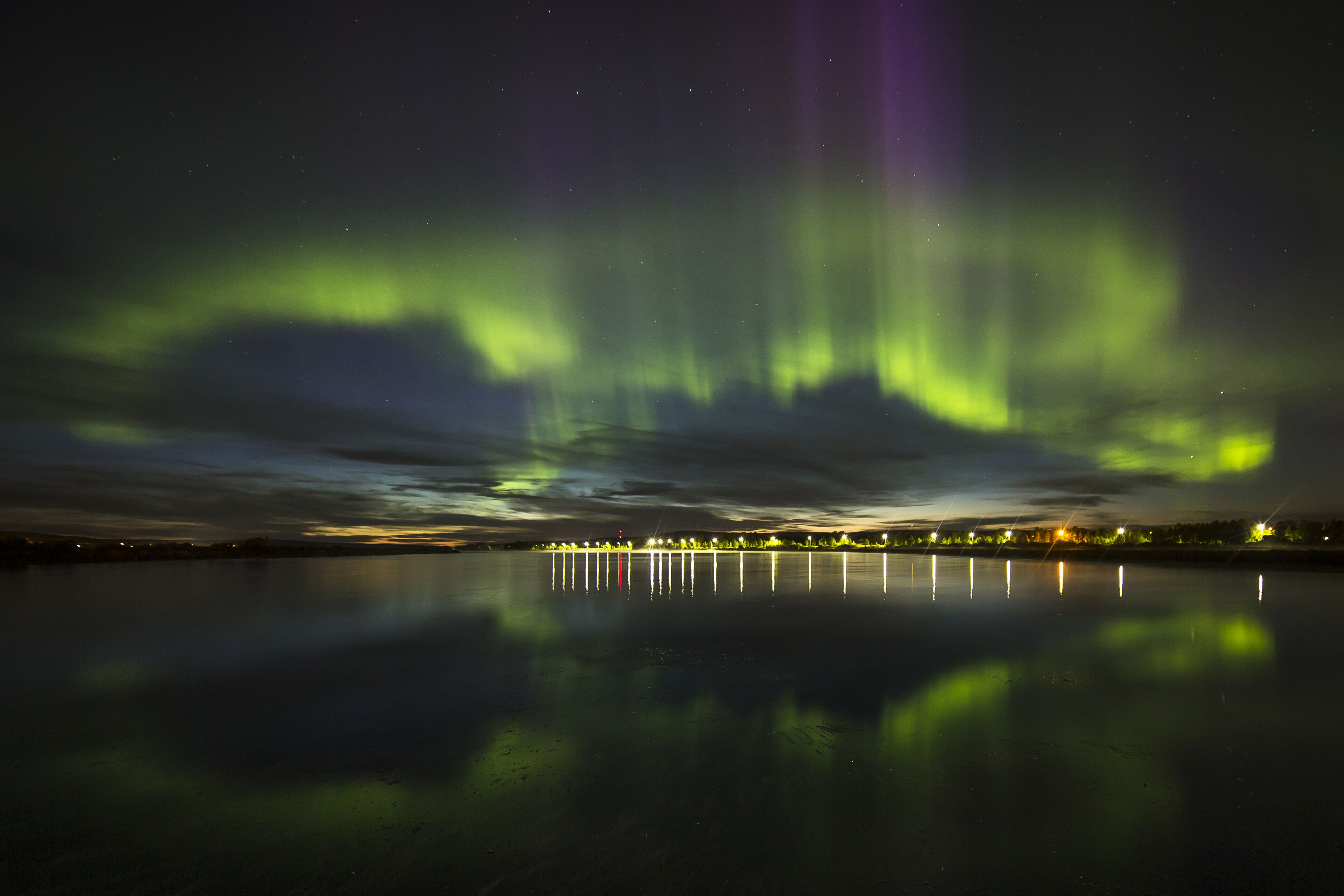 Una escena nocturna con las auroras boreales reflejadas en aguas tranquilas, con un horizonte urbano de fondo.