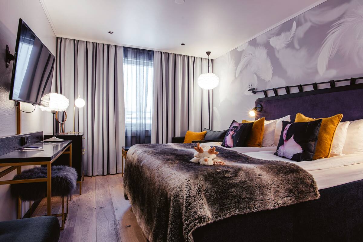A modern hotel room with a large bed, a desk with a computer, a TV, and a large window with curtains.