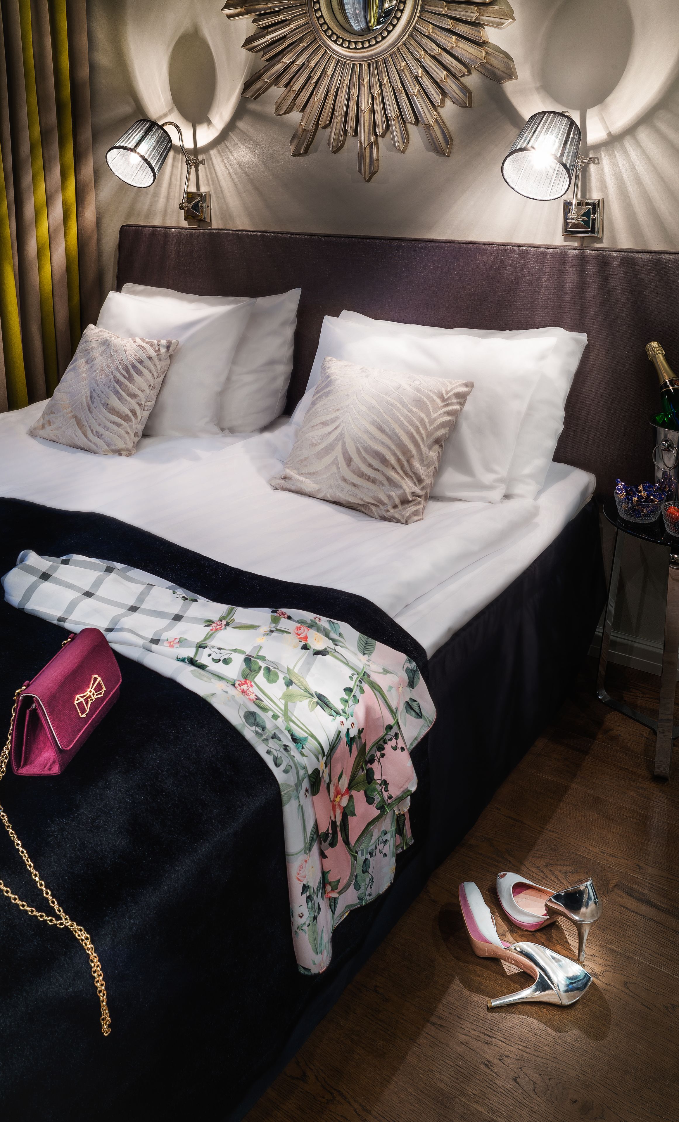 Una cama bien hecha con sábanas blancas y almohadones decorativos, una manta con estampado floral, un bolso rosa y un par de zapatos de tacón en el suelo.