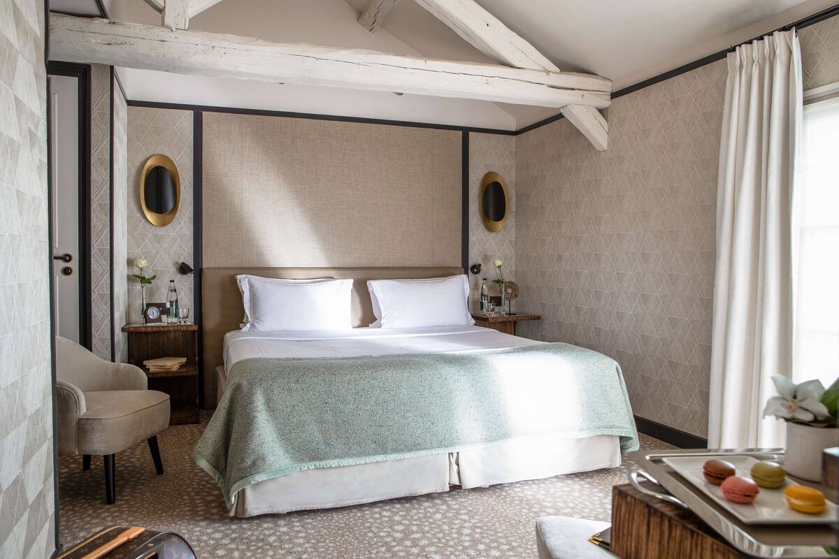 A neatly arranged bedroom with a bed, a chair, a bedside table with a lamp, and a tray with macarons.