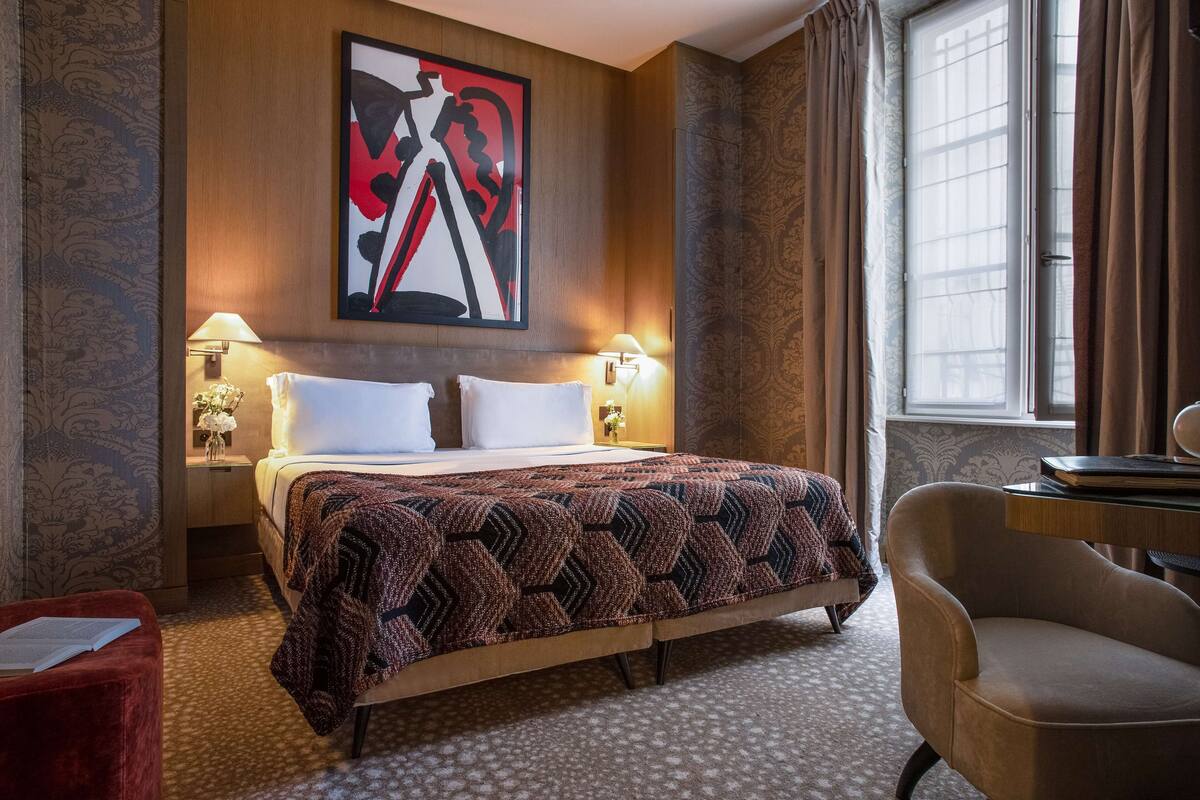 A hotel room with a large bed, two bedside lamps, a desk with a chair, a window with curtains, and a wall with a large abstract painting.