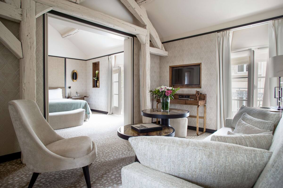 A modern hotel room with a large bed, a sitting area with a sofa and armchair, a small table with a vase of flowers, and a TV mounted on the wall.