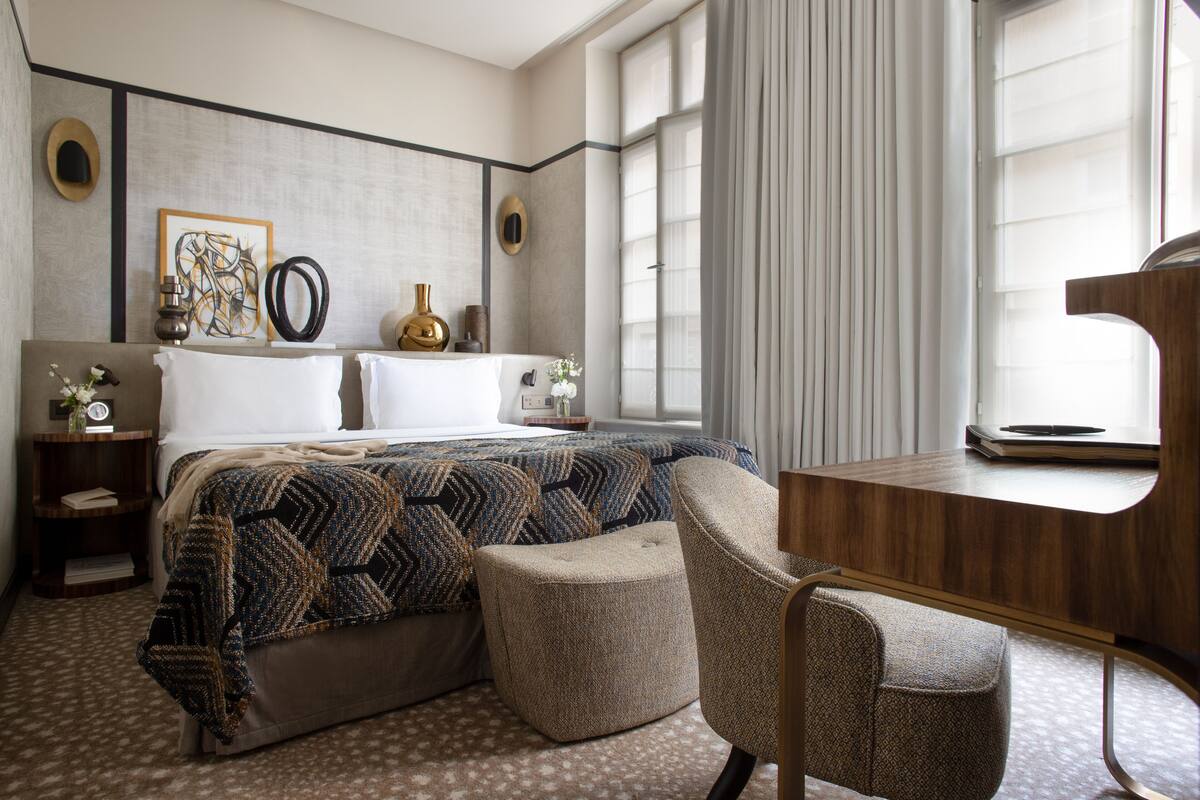 A hotel room with a bed, a desk, and a chair. There are two bedside tables with lamps and a clock. The room has a window with curtains.