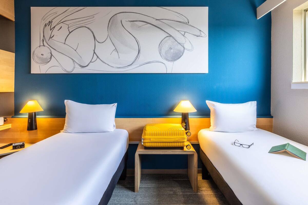 Une chambre d’hôtel avec deux lits, un mur d’accent bleu et un grand tableau abstrait.