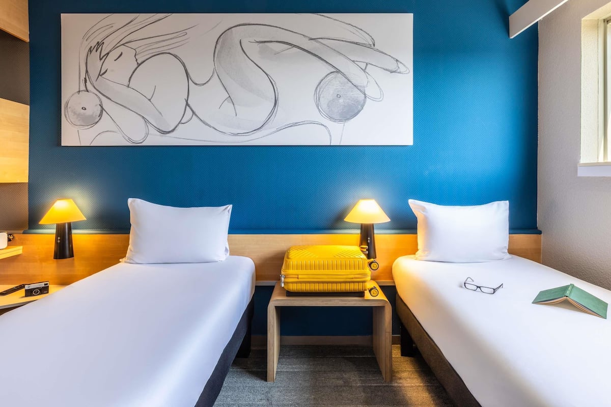 Une chambre d’hôtel avec deux lits, un mur d’accent bleu et un grand tableau abstrait.