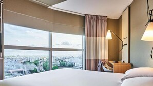 A hotel room with a large bed, a desk, a chair, a lamp, and a view of a cityscape through large windows. - ibis Paris Porte De Montreuil (Paris)