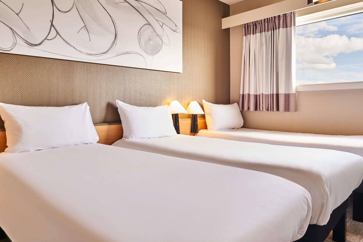 Une chambre d’hôtel avec deux lits, une grande peinture abstraite au mur, une fenêtre avec des rideaux et une lampe sur la table de chevet.