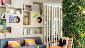 A modern living room with a sofa, coffee table, and a chair. There are bookshelves with books and decorative items. A green wall with plants is visible. - ibis Paris Porte De Montreuil (Paris)