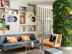 Ein modernes Wohnzimmer mit einer Couch, einem Couchtisch und einem Sessel. Bücherregale mit Büchern und Dekorationen sind zu sehen. Eine grüne Wand mit Pflanzen ist ebenfalls erkennbar.