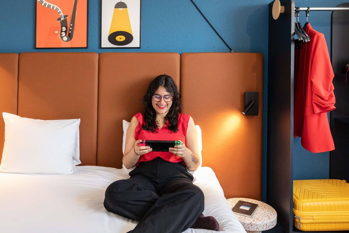 Une personne est assise sur un lit, tenant une tablette. Sur le mur, on aperçoit un cadre avec une photo de saxophone et une ampoule. Un sac de voyage jaune est posé sur le sol.