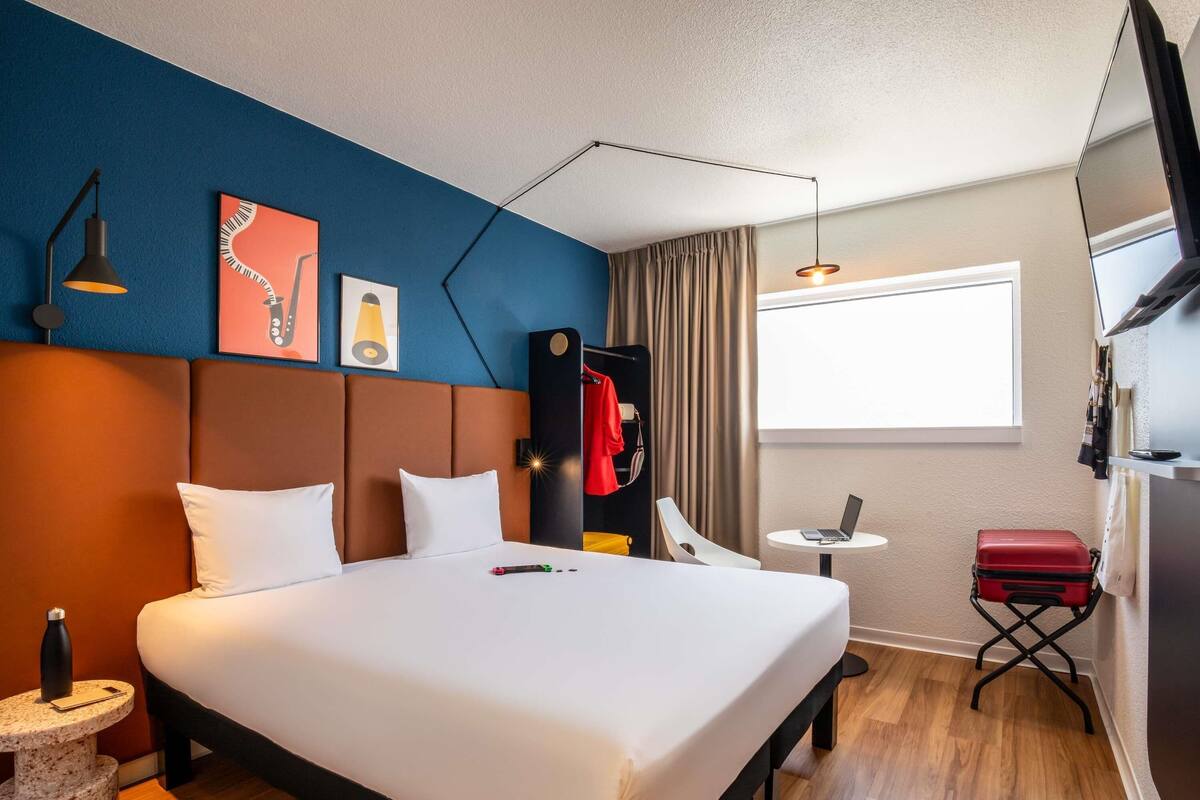 Une chambre d’hôtel avec un lit, un bureau avec un ordinateur portable, une télévision et une valise.