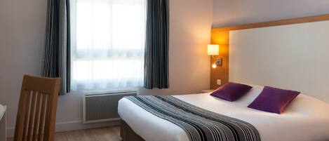 A hotel room with a bed, a chair, a window with curtains, a wall-mounted lamp, and a radiator.