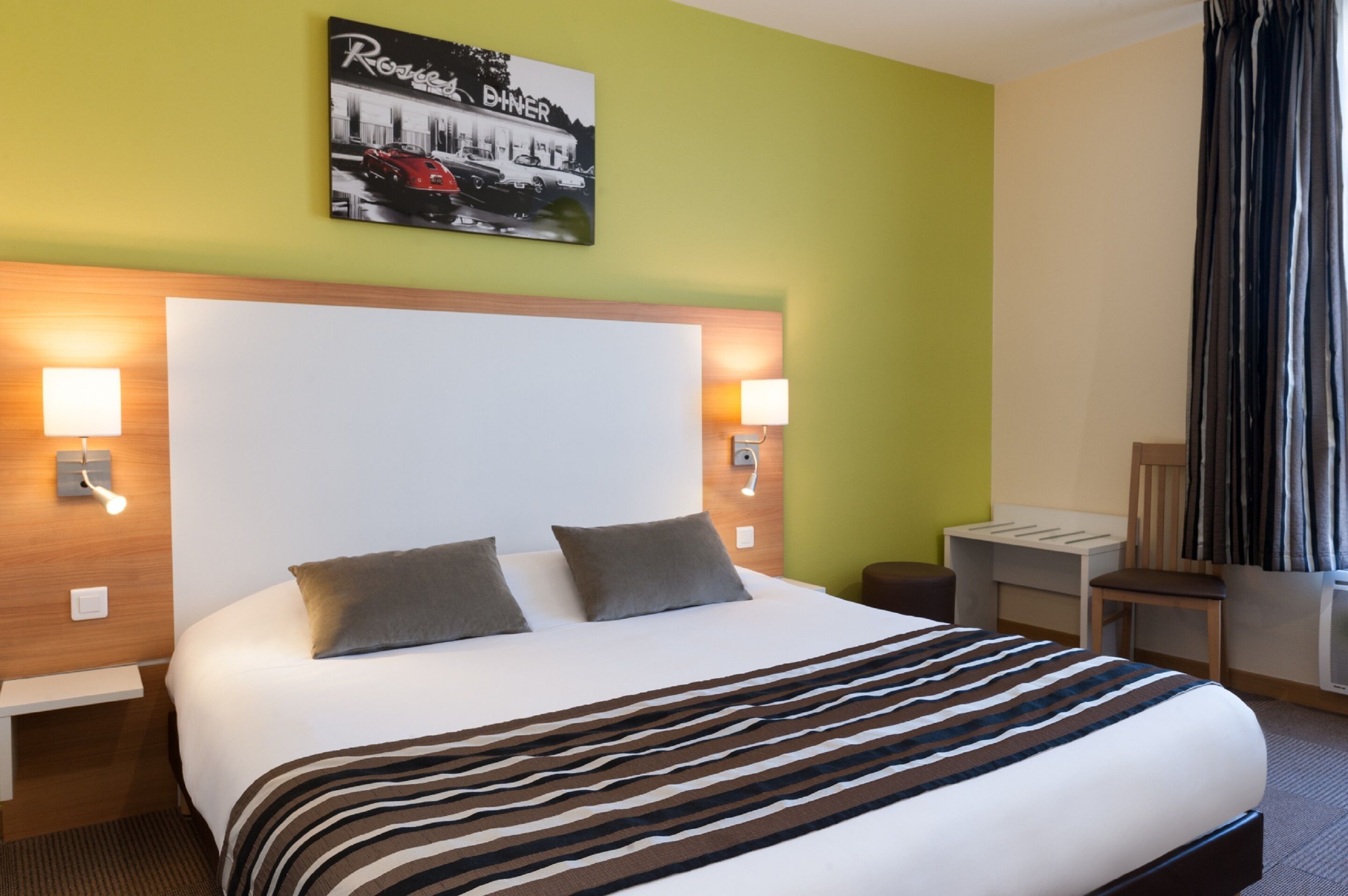 Habitación de hotel con una cama grande, dos lámparas de noche, cabecera, una mesita pequeña, una silla y un ventanal con cortinas.