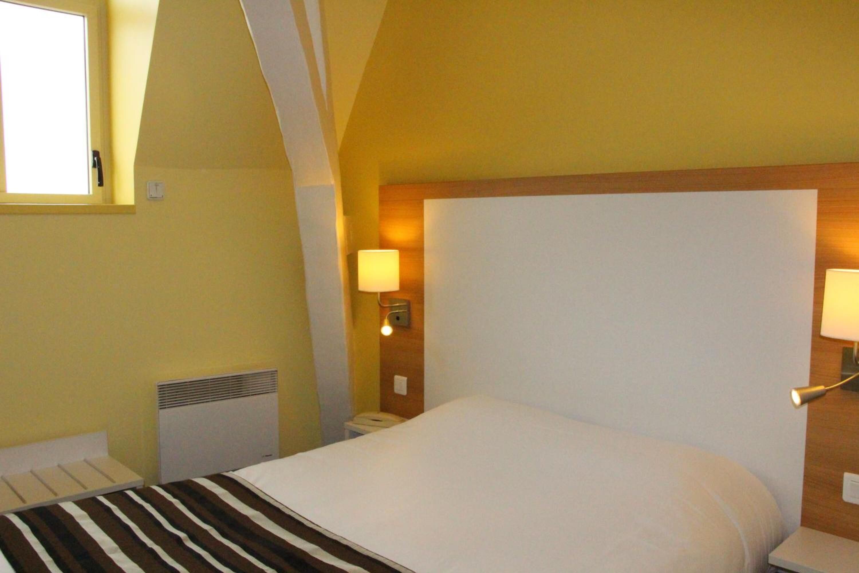 A hotel room with a bed, a bedside table, a lamp, and a window.