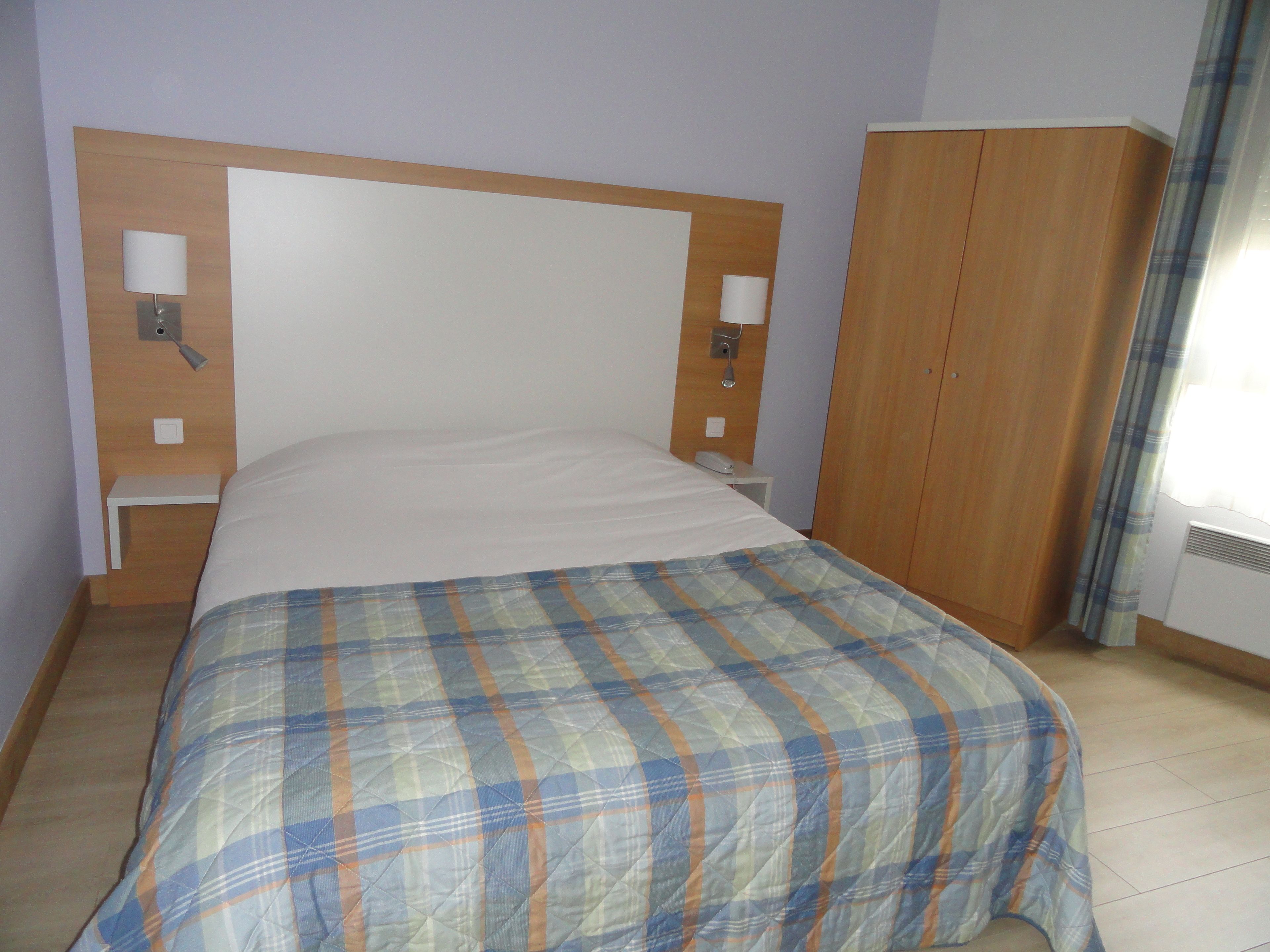 Una habitación de hotel con una cama, cabecera de madera, dos lámparas de noche, una mesita de noche y un armario.