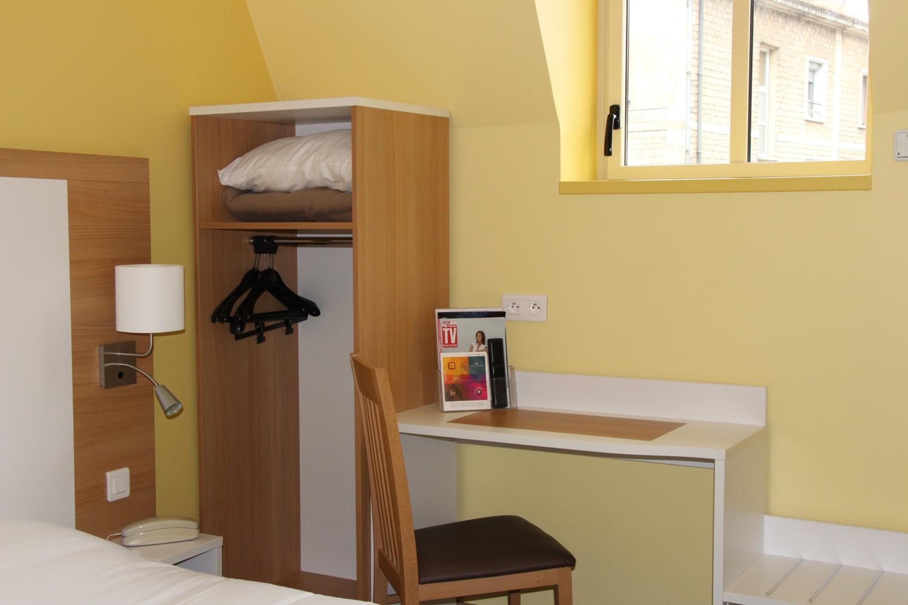 Habitación de hotel pequeña con cama, escritorio con silla, armario, televisión y ventana con vistas a edificios.