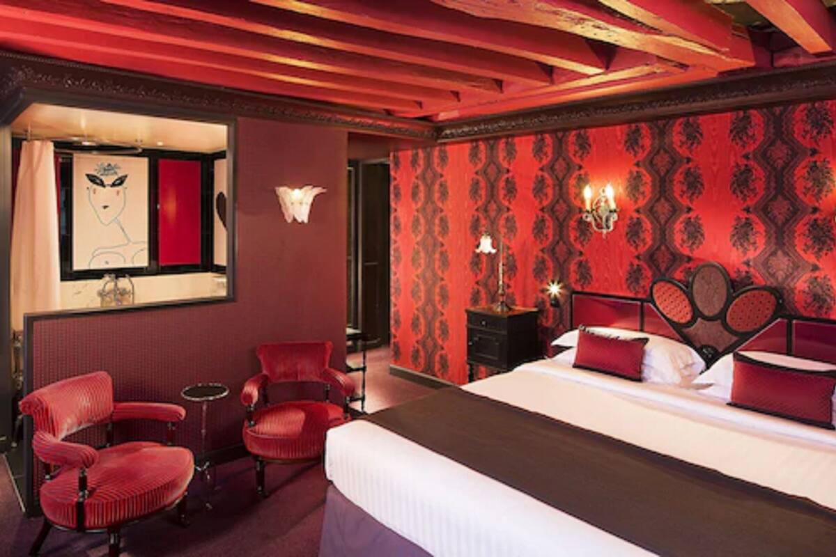 A hotel room with a large bed, red upholstered chairs, a mirror, and a wall with a patterned wallpaper.