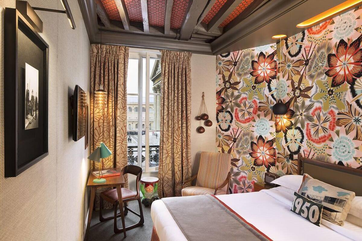 A hotel room with a bed, a desk with a lamp, a chair, a window with curtains, and a wall with a floral pattern.