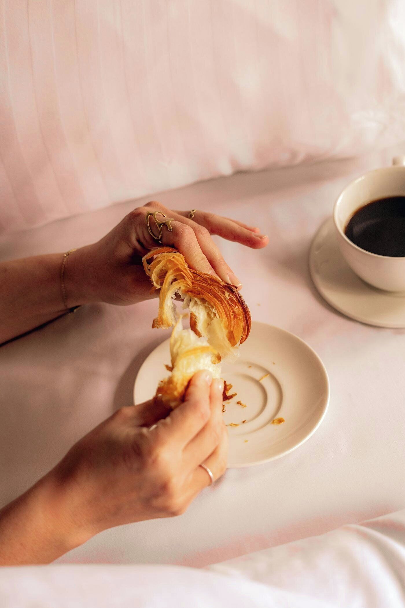 A person holding a piece of bread with a bite taken out of it, a plate with crumbs, and a cup of coffee on a table.