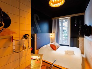 Une chambre d’hôtel avec un lit, une fenêtre, une étagère fixée au mur et un luminaire suspendu.