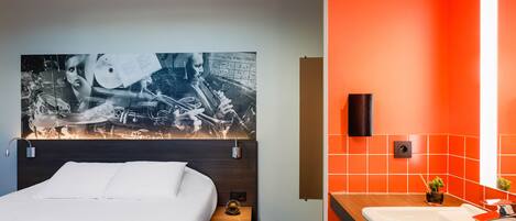 Ein Hotelzimmer mit einem großen Bett, einem Kopfteil mit einem Schwarz-Weiß-Foto, einem kleinen Nachttisch mit einer Lampe, einem modernen Badezimmer mit einem großen Spiegel und orangefarbenen Wänden.