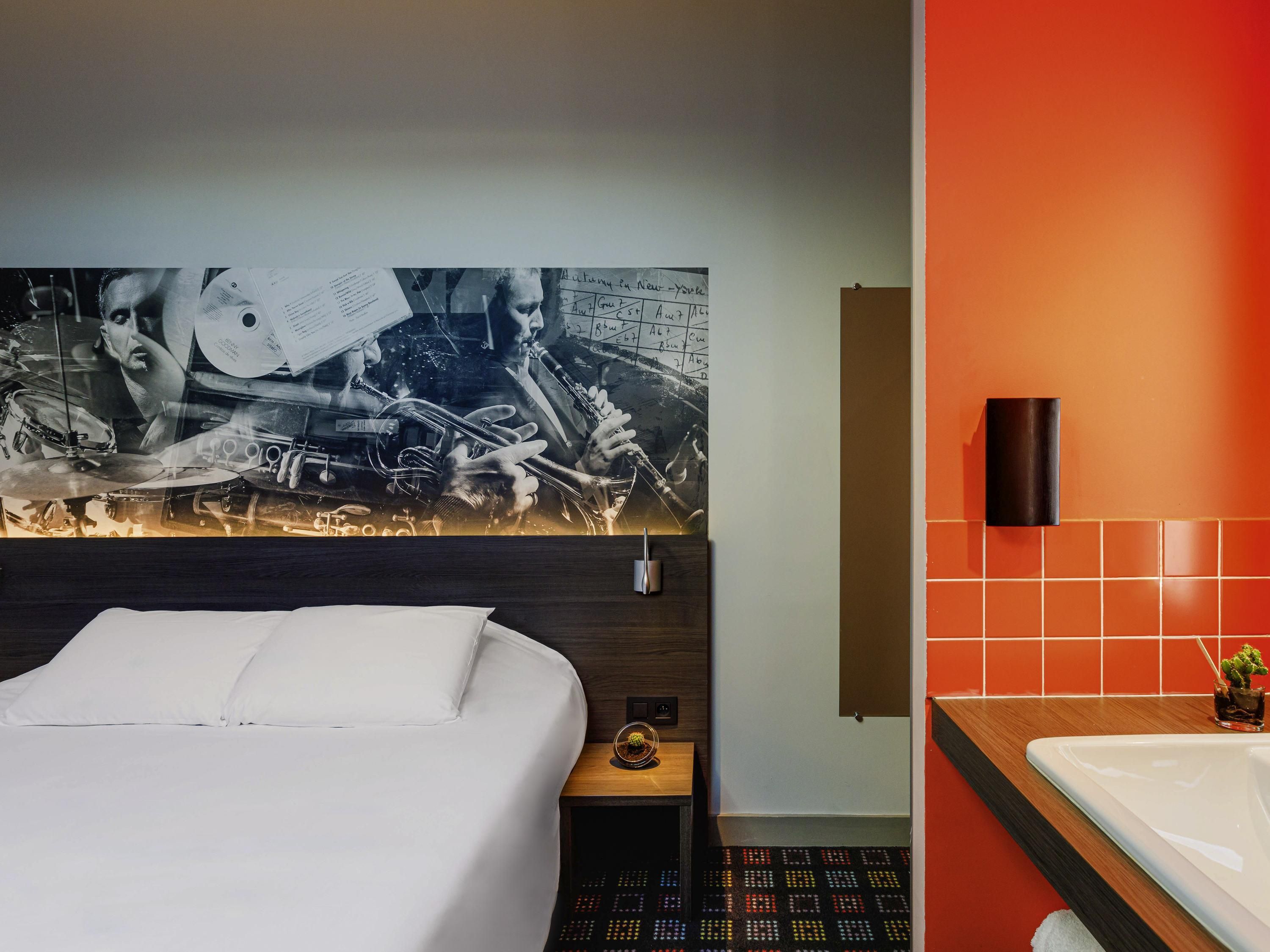 Habitación de hotel con un cartel grande de músicos, una cama con sábanas blancas, una mesita de noche pequeña con un reloj y un baño con pared de azulejos rojos y lavamanos blanco.
