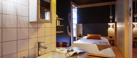 Habitació d'hotel moderna amb un ampli tocador al bany, un llit amb capçalera de fusta i una taula petita amb un llibre i uns gots.