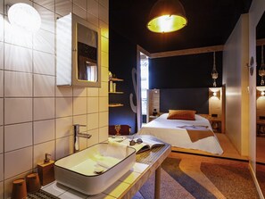 Camera d'albergo moderna con un ampio lavabo in bagno, un letto con testiera in legno e un piccolo tavolino con un libro e dei bicchieri.