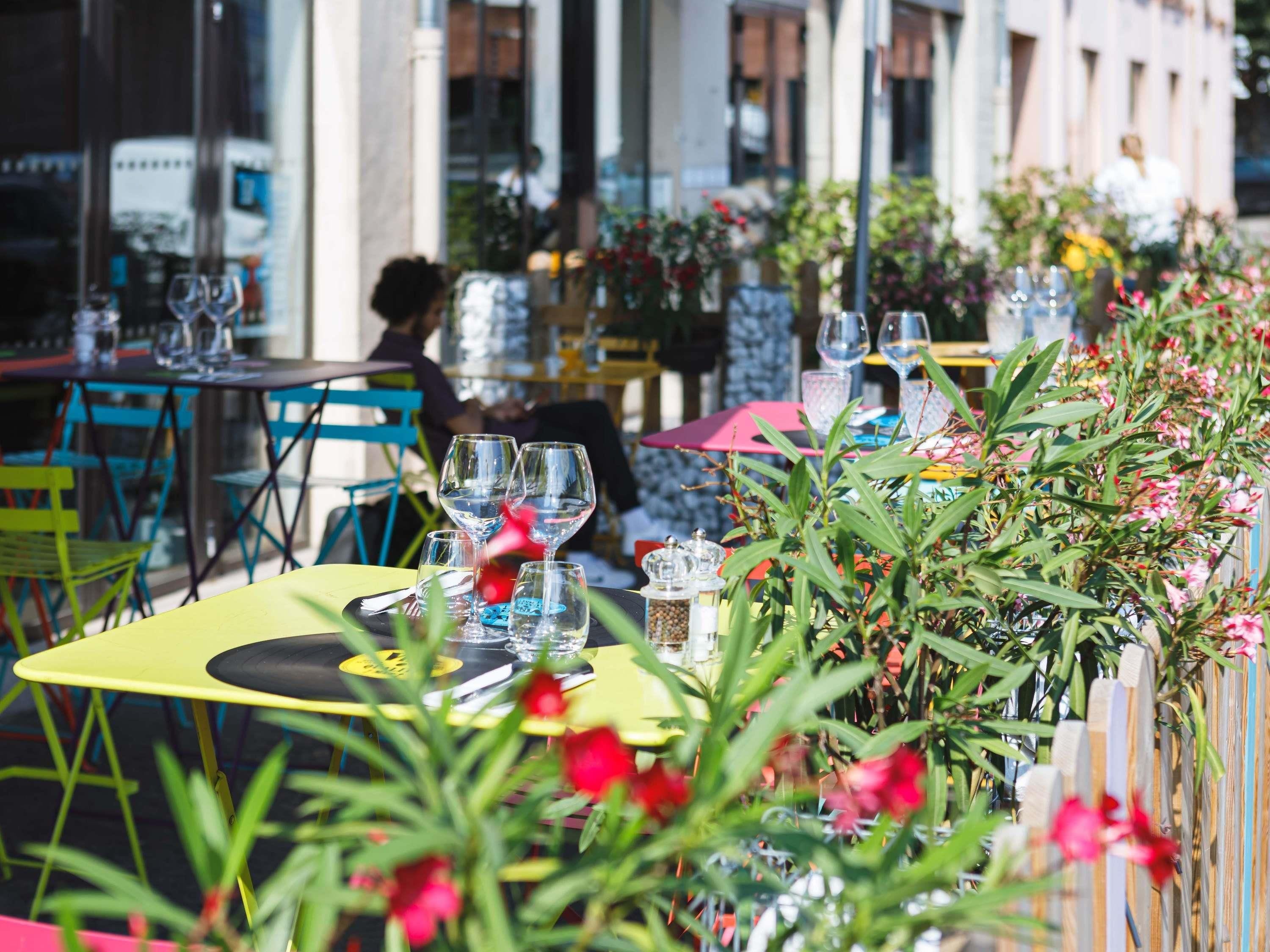 An outdoor cafe with tables and chairs, a person seated, and potted plants.