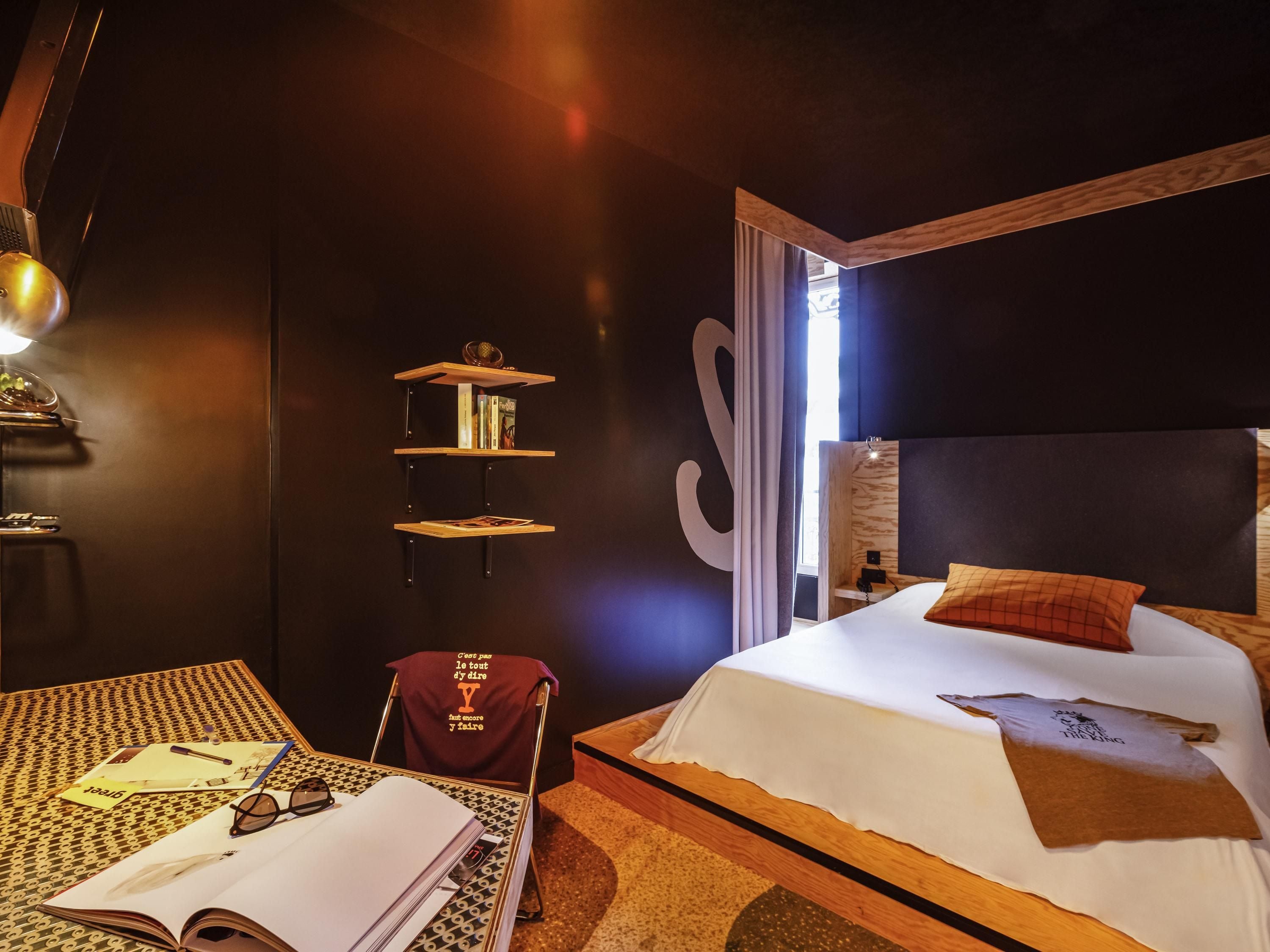 Habitación de hotel con cama, escritorio, silla, estantería y un amplio ventanal.