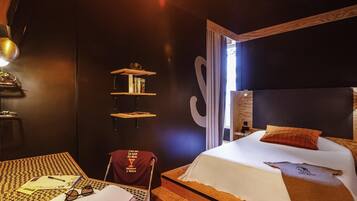 Habitación de hotel con cama, escritorio, silla, estantería y un amplio ventanal.