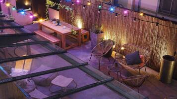 Una terraza en la azotea con una mesa de madera, sillas de mimbre y luces colgantes.