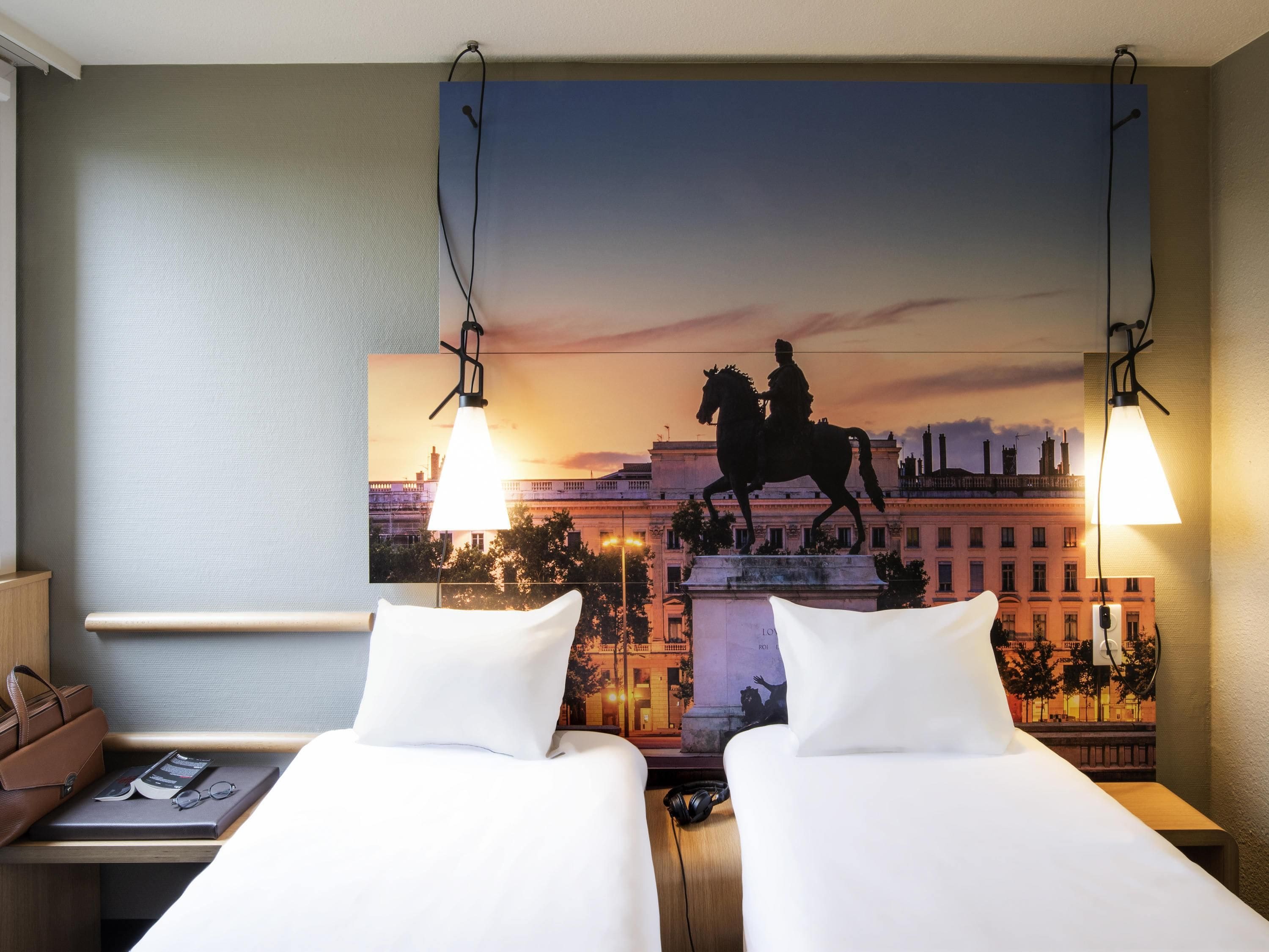 Een hotelkamer met twee bedden, een nachtkastje, een lamp, een schilderij van een paardenbeeld en uitzicht op een stadslandschap.