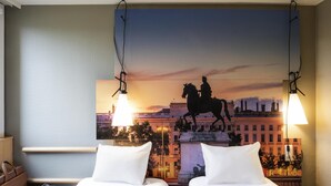 A hotel room with two beds, a nightstand, a lamp, a painting of a horse statue, and a cityscape view. - ibis Lyon Gerland Rue Merieux (Lyon)