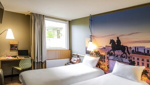 A hotel room with two beds, a desk, a chair, a lamp, and a large cityscape mural on the wall. - ibis Lyon Gerland Rue Merieux (Lyon)