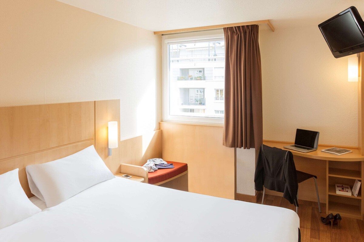 Habitación de hotel con cama, escritorio con computadora portátil, silla, televisión y ventana con cortinas.