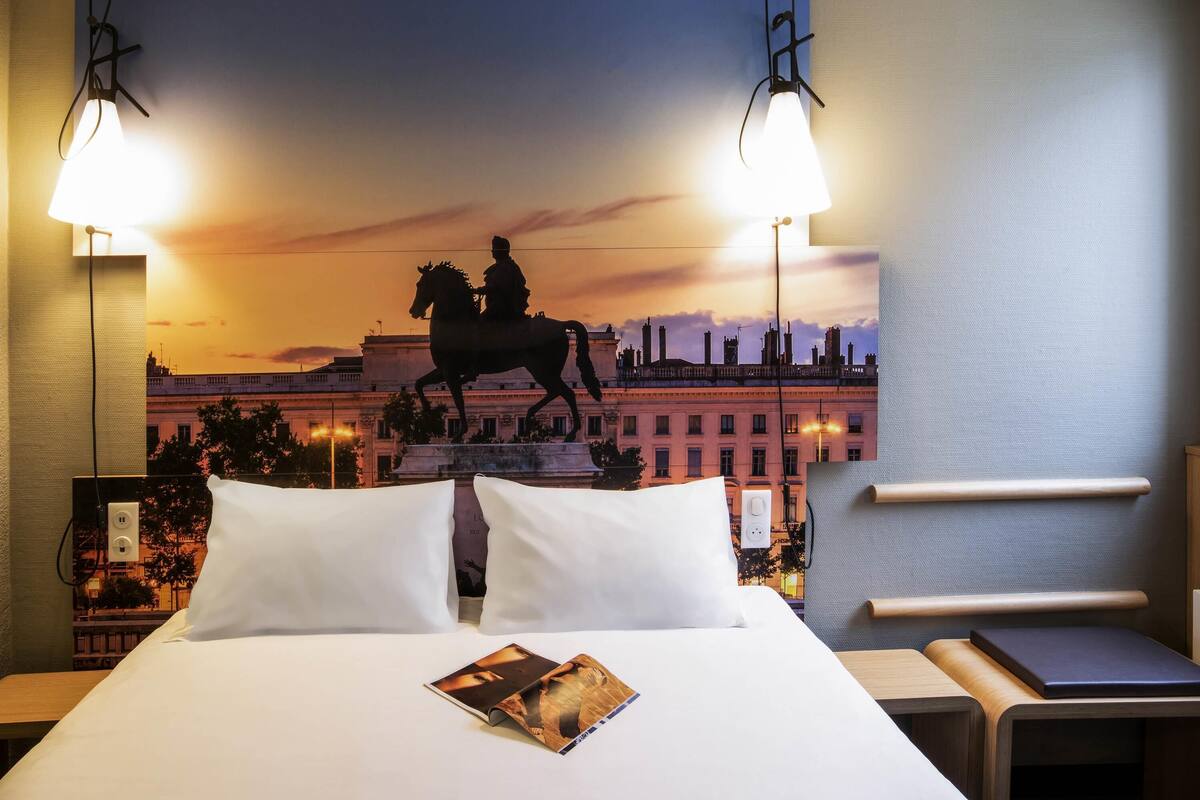 A hotel room with a bed, a nightstand, a reading light, and a wall-mounted lamp.