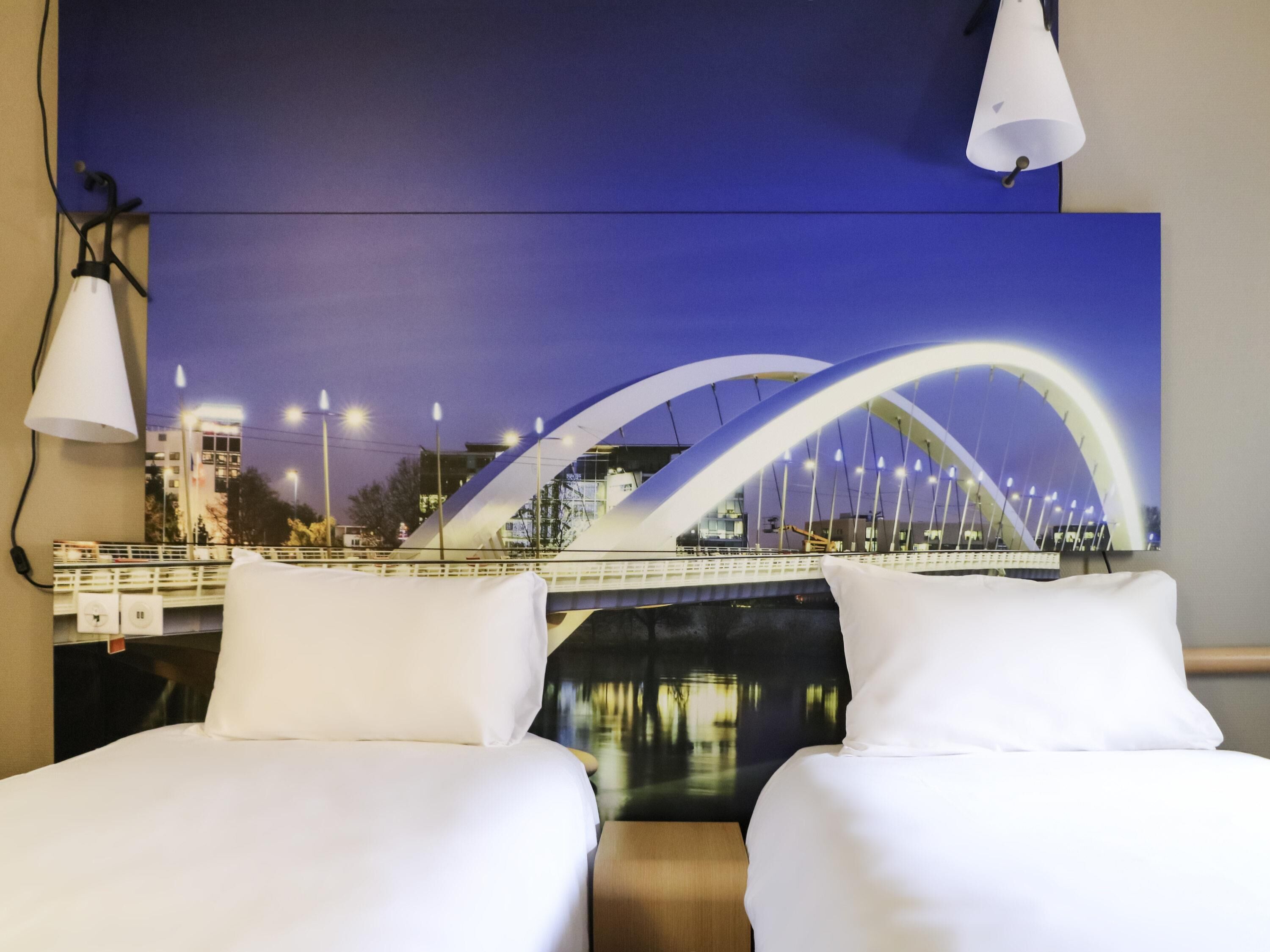 A hotel room with two beds, a large wall-mounted TV displaying a cityscape, and a view of a bridge.