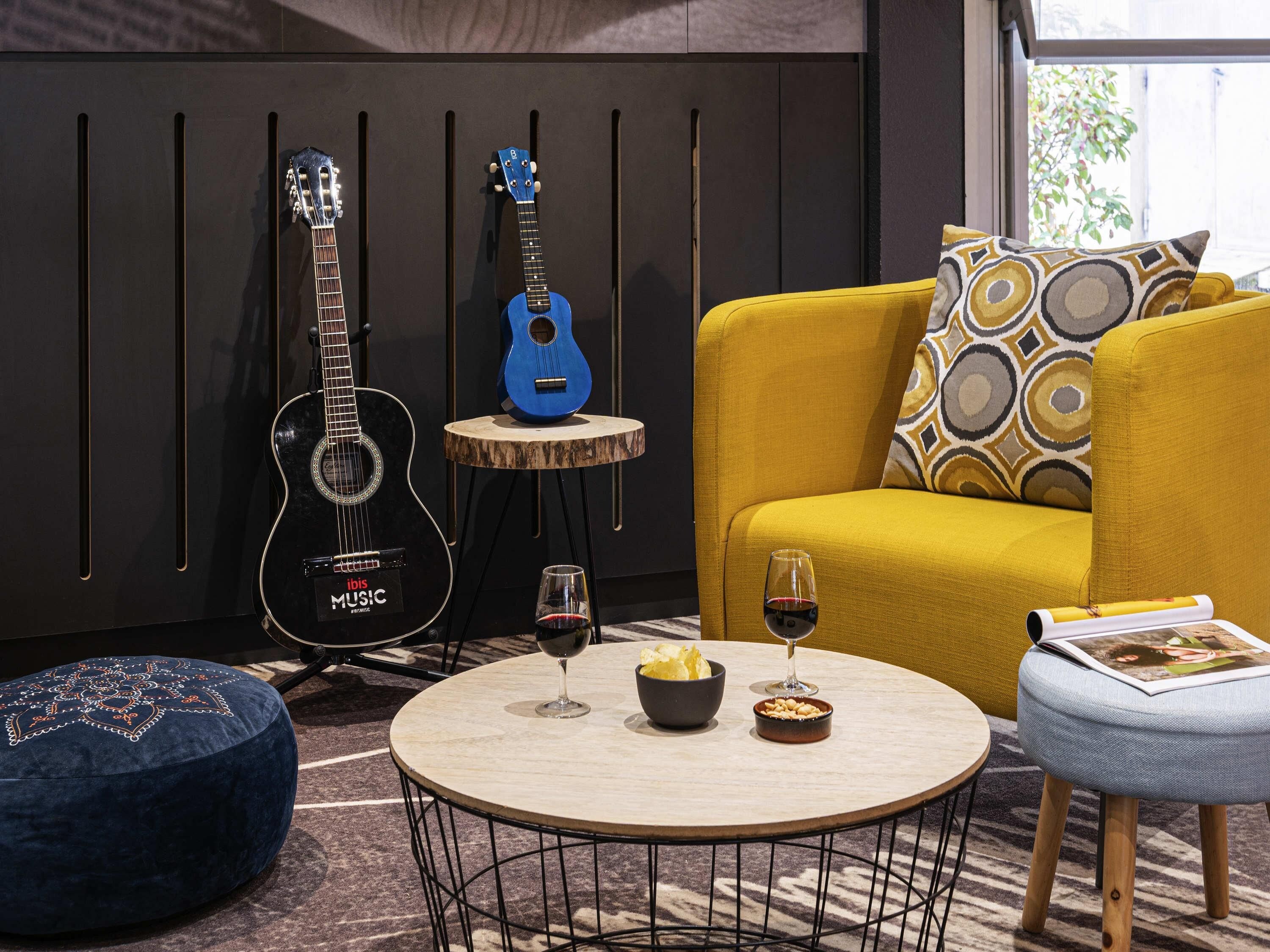 Een kamer met een gele bank, een blauwe gitaar, een ronde tafel met wijnglazen en een blauwe poef.